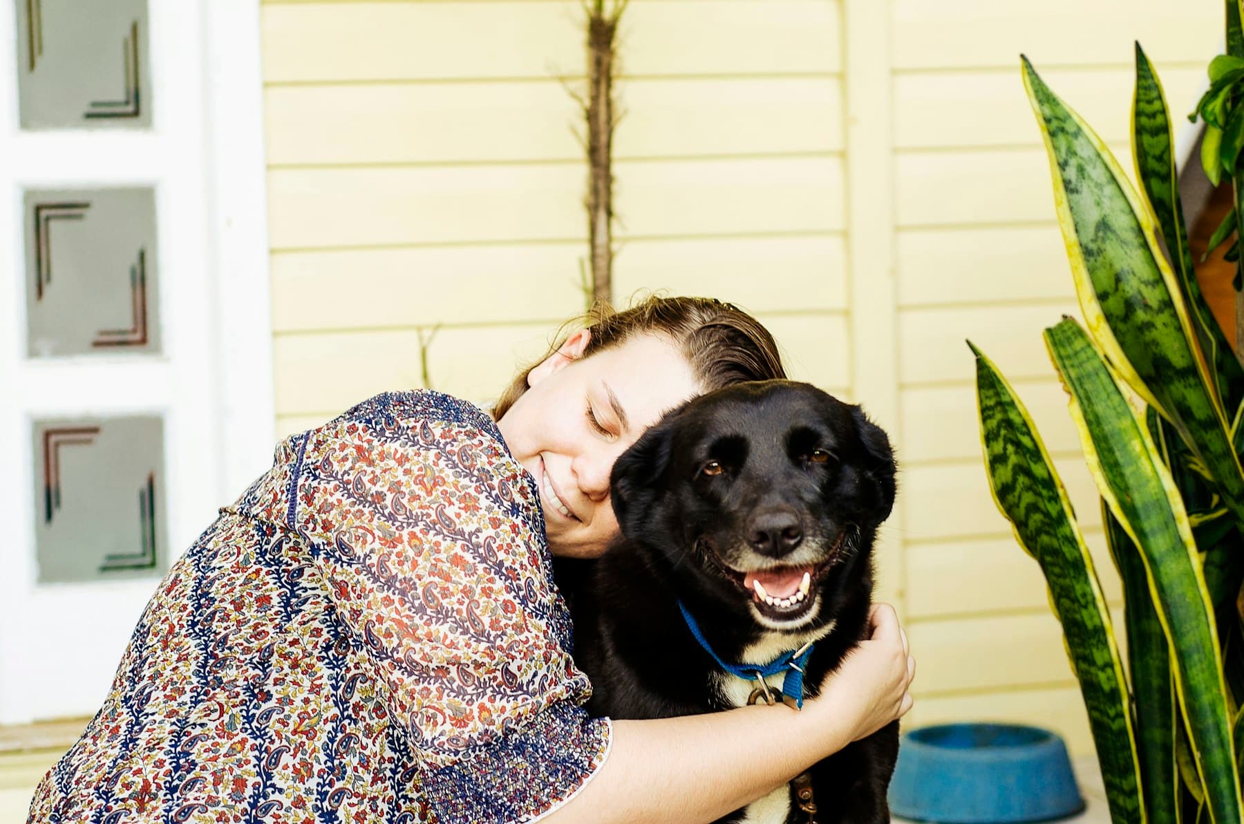 A woman with her eyes closed, smiling as she cuddles her happy black dog on the porch