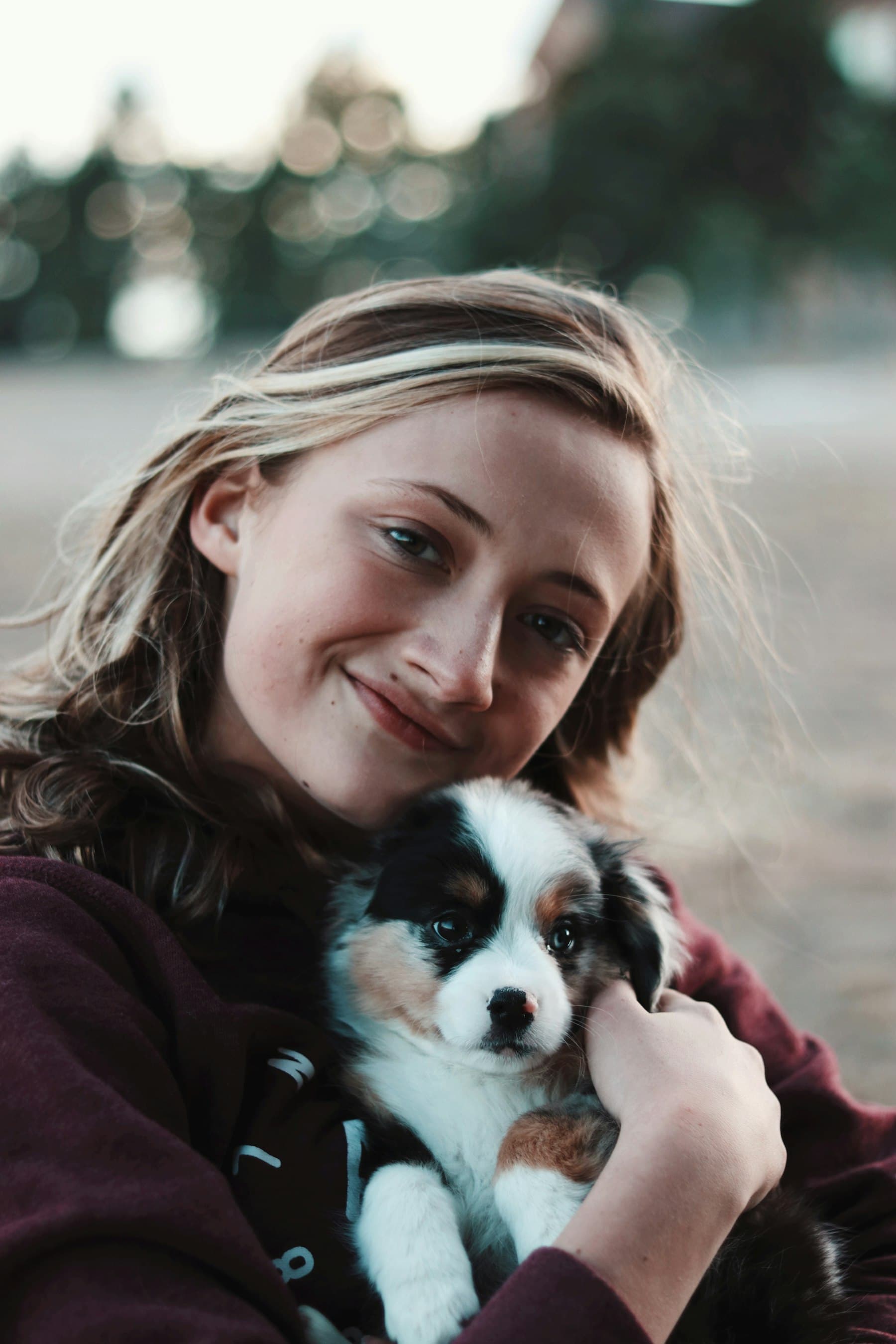 A smiling young woman holding a fluffy tricolour puppy close to her cheek outdoors