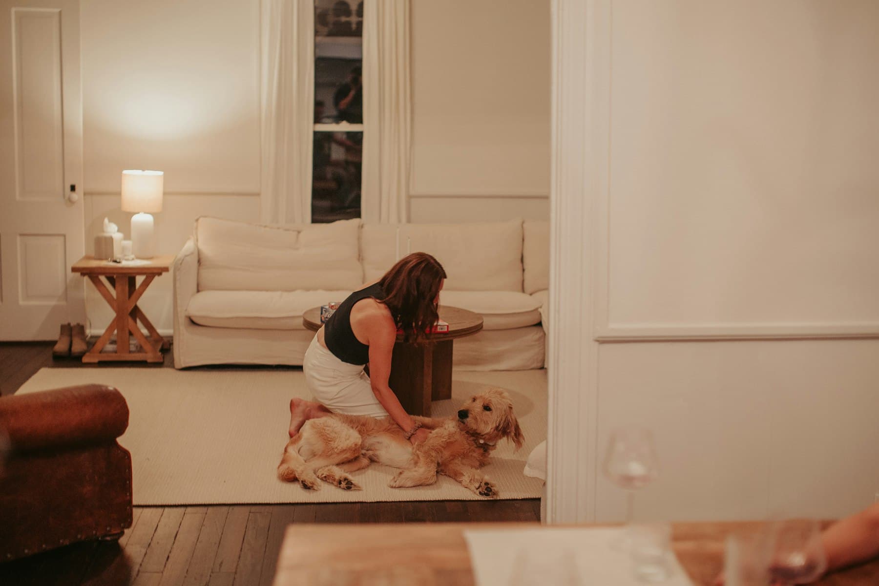 A woman kneeling on a rug in a warmly lit living room, stroking her relaxed golden doodle